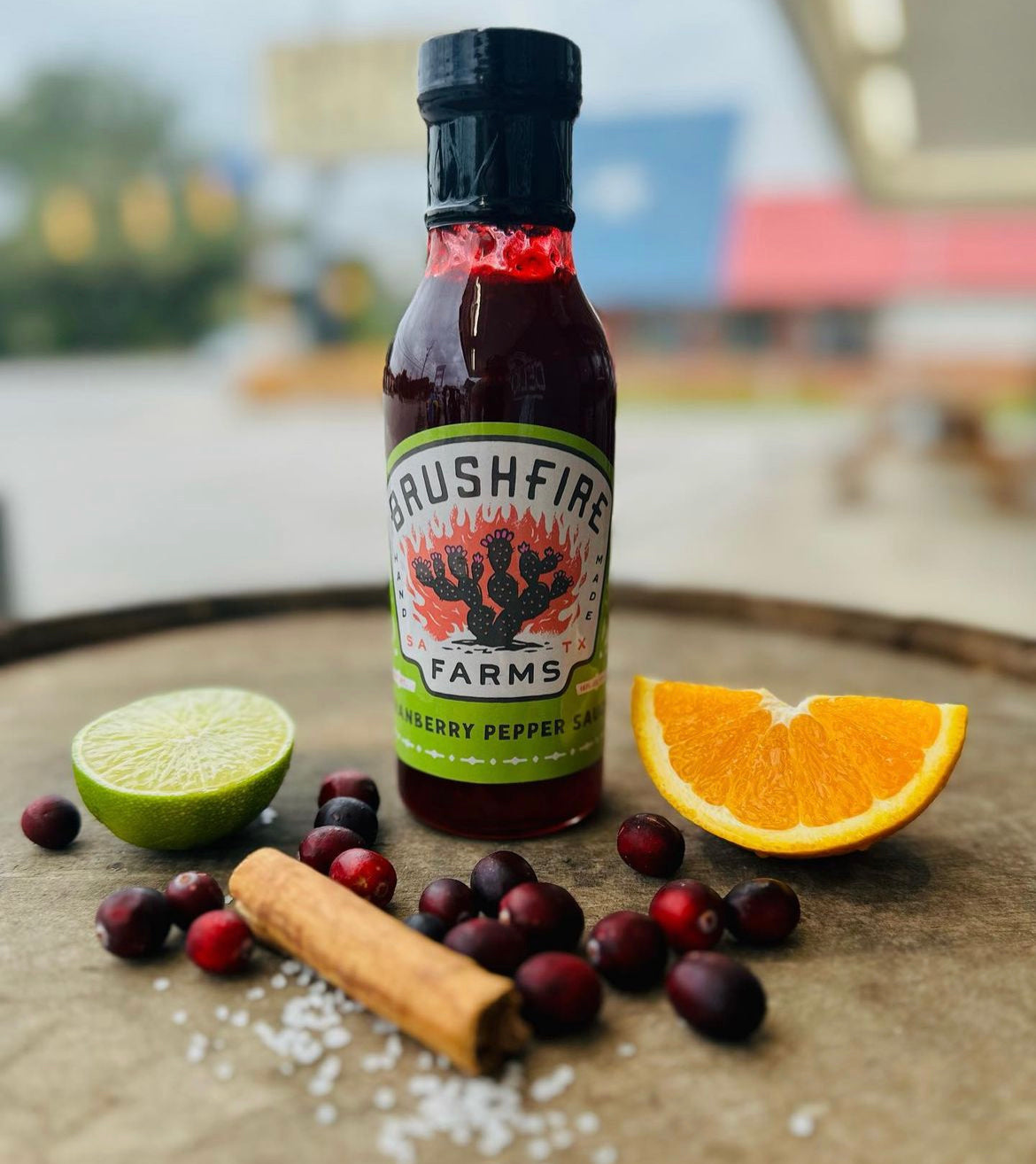 Cranberry Pepper Sauce bottle with lime, orange slices, cranberries, cinnamon stick, and salt on a wooden surface.