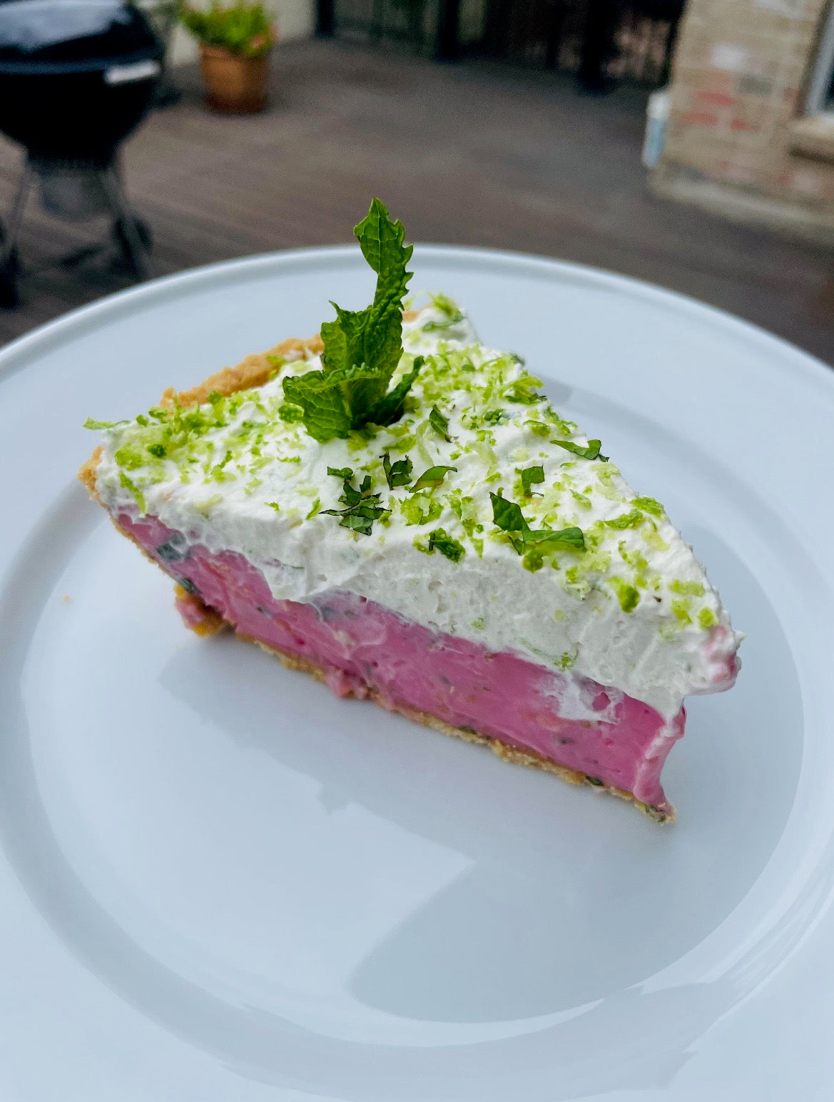 Slice of prickly pear pie topped with whipped cream, lime zest, and mint on a white plate, showcasing vibrant pink filling.