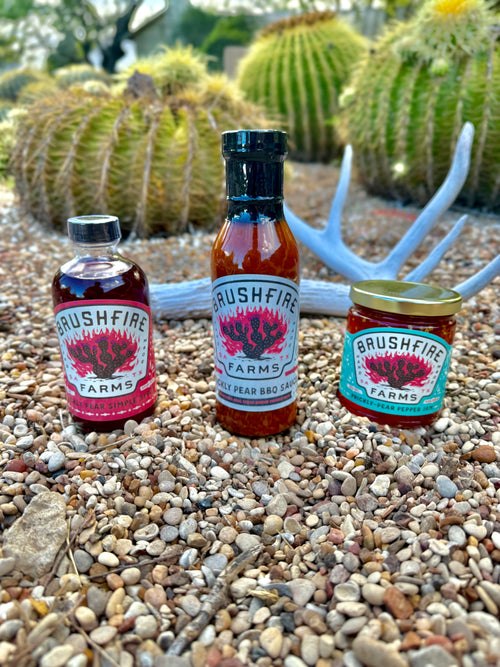 Three jars of Brushfire Farms' Prickly Pear Pack featuring prickly pear syrup, BBQ sauce, and jam against a desert backdrop.