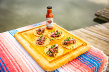 Load image into Gallery viewer, Bottle of Brushfire Farms Prickly Pear BBQ sauce on a wooden cutting board with steak fajitas on a colorful towel by a body of water.