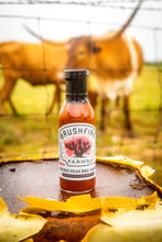 Load image into Gallery viewer, Bottle of Brushfire Farms prickly pear barbecue sauce on a wooden surface with a Longhorn in the background.