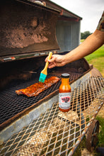 Load image into Gallery viewer, Person grilling ribs with a bottle of Brushfire Farms Prickly Pear BBQ sauce on a grill outdoors.