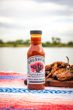 Load image into Gallery viewer, Bottle of Brushfire Farms prickly pear BBQ sauce on a colorful blanket with grilled food in the background.