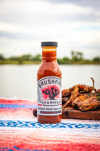 Bottle of Brushfire Farms prickly pear BBQ sauce on a colorful blanket with grilled food in the background.